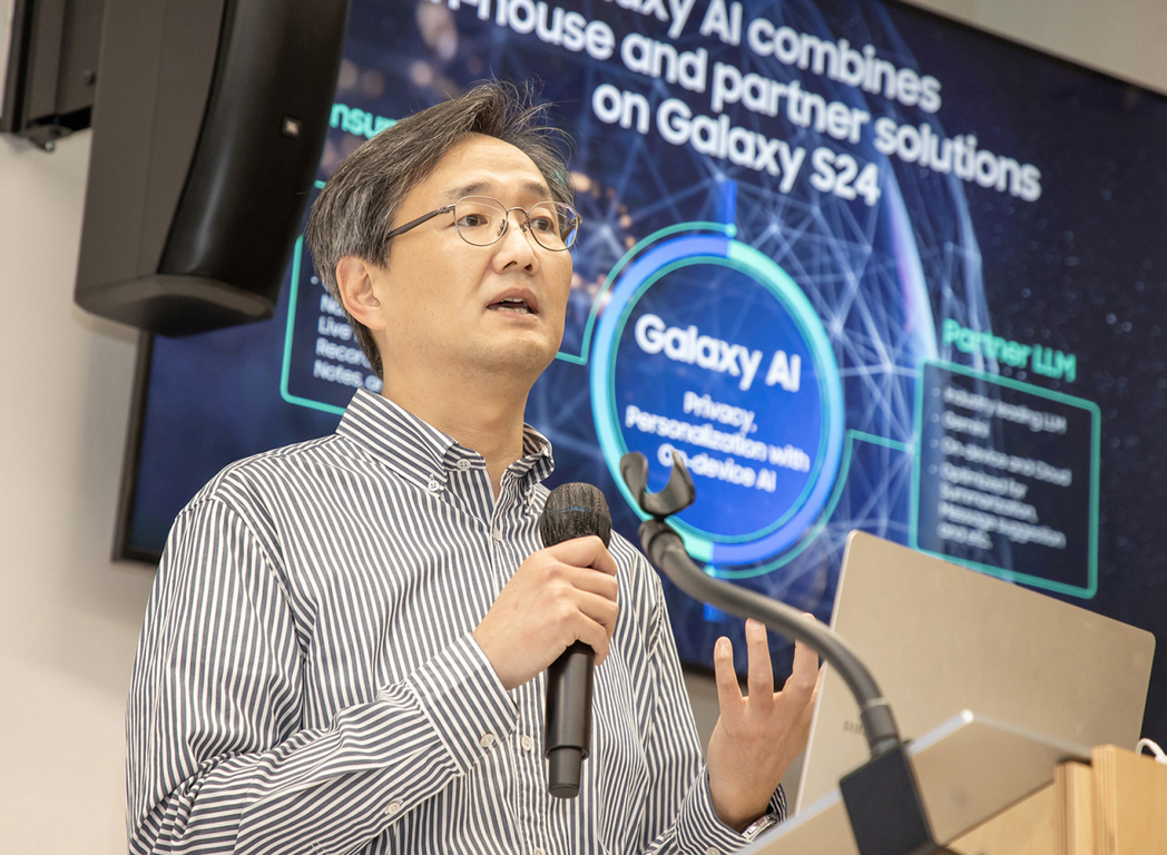안경을 쓴 남성이 줄무늬 셔츠를 입고 마이크를 들고 발표하고 있다. 뒤에는 'Galaxy AI'와 'Galaxy S24' 관련 정보가 적힌 대형 스크린이 보인다.