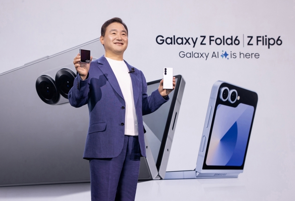 남성이 무대에서 갤럭시 Z 폴드6와 Z 플립6 스마트폰을 각각 한 손에 들고 소개하고 있으며, 배경에는 제품 이미지와 'Galaxy Z Fold6 | Z Flip6 Galaxy AI is here'라는 문구가 보인다.