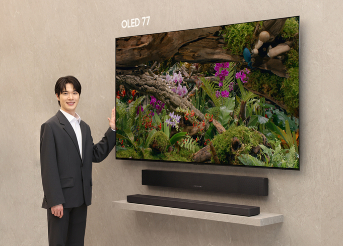 정장 차림의 남성이 벽에 설치된 77인치 OLED TV 옆에 서서 미소 짓고 있다. TV 화면에는 자연 풍경이 생생하게 표현되어 있고, 아래에는 사운드바가 놓여 있다.