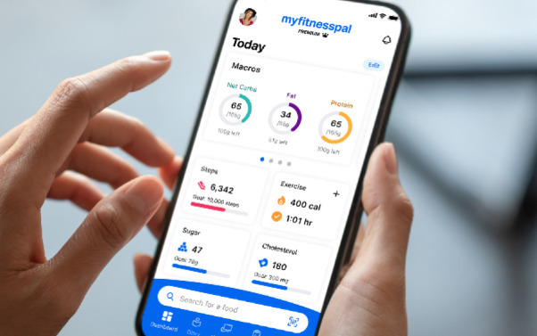 한 사람이 손에 스마트폰을 들고 MyFitnessPal 건강 관리 앱의 대시보드를 보고 있다. 화면에는 영양소 섭취량, 걸음 수, 운동 칼로리 소모 등 건강 관련 정보가 표시되어 있다.