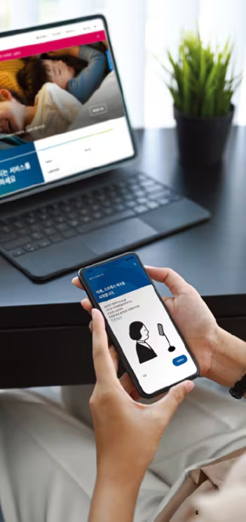 모바일 AI 건강관리 앱 활용법: 건강 트래킹부터 데이터 보안까지 2 사람이 책상에 앉아 노트북과 스마트폰을 사용하고 있으며, 노트북에는 가족 사진이 있는 웹사이트가 열려 있고, 스마트폰에는 여성의 음성 인식 관련 화면이 표시되어 있다. 배경에는 작은 화분이 놓여 있다.